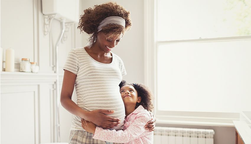 A pregnant woman hugs her belly while hugging her daughter with her other arm. The young daughter has both arms wrapped around her mom's waist.. 